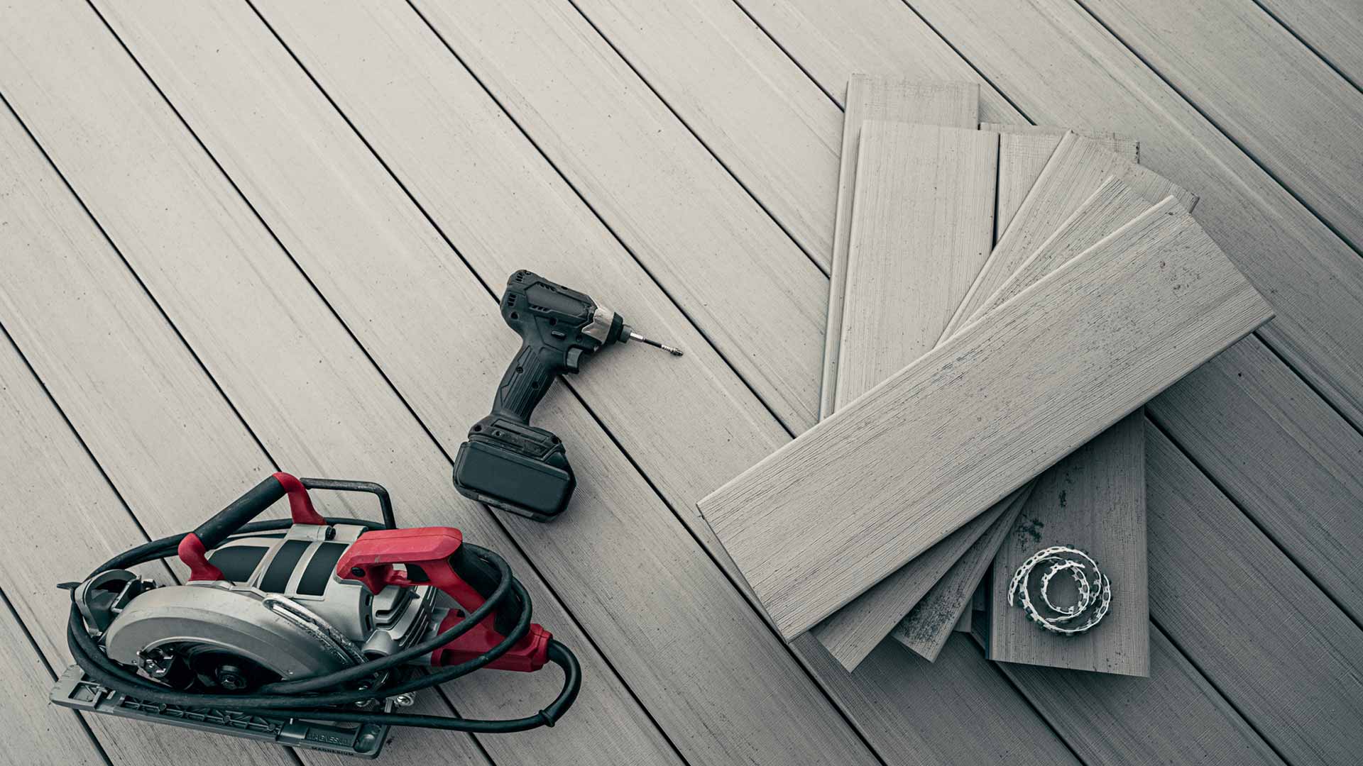 A drill, circular saw, and deck board pieces on a gray deck