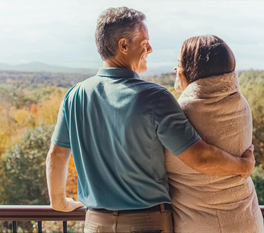 Lori is wrapped in a blanket and is standing next to Mike, who wraps his arm around her. The two have big grins on their face and are facing the mountain view from their deck.