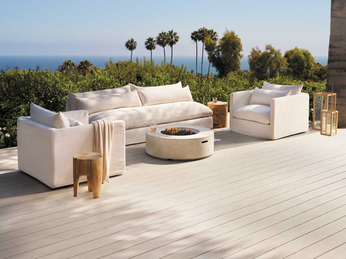 Modern, cushy white linen armchairs and a couch on a light tan deck with the beach in the background