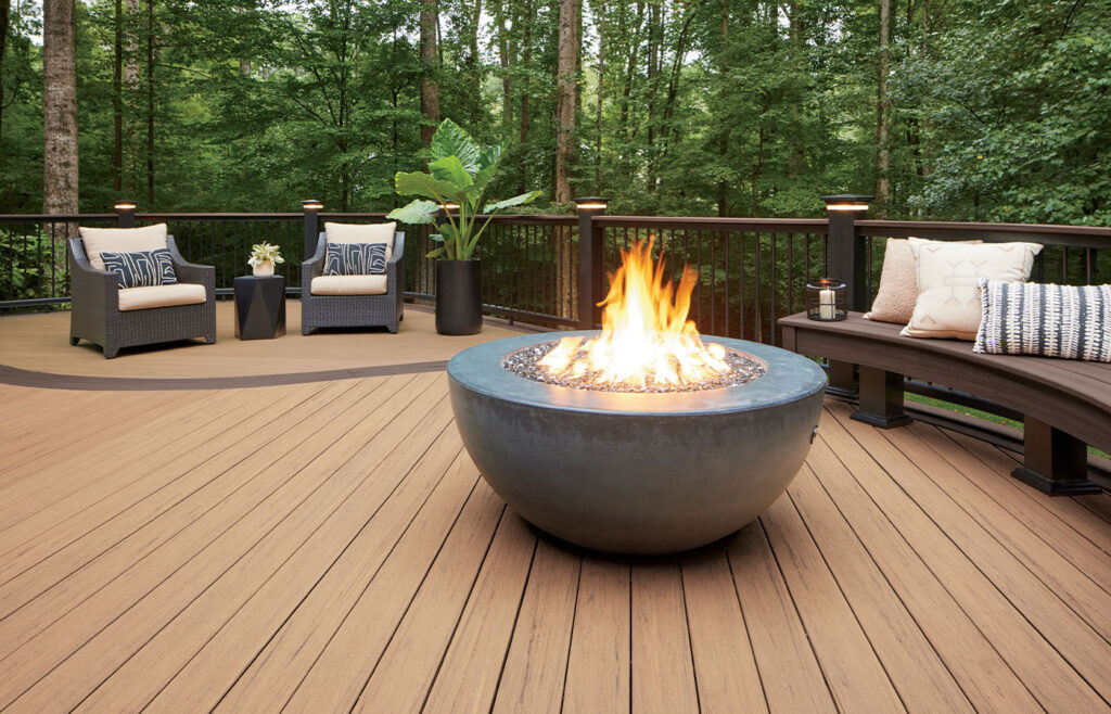 A wooden deck surrounded by trees features two armchairs, a bench, and a large, lit fire pit in a round bowl.