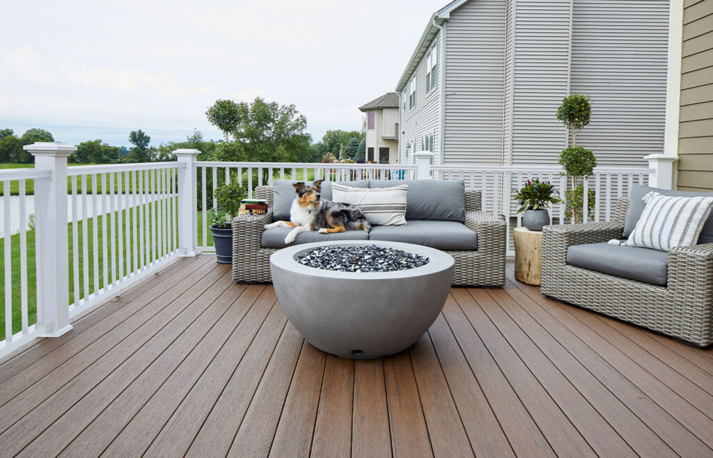 A deck trimmed with white railing from our Radiance Rail Express line and dark brown Dark Roast decking from our Reserve Collection, during dusk. The deck is decorated with a light gray fire pit and light gray wicker furniture. A dog sits on a sofa behind the fire pit.
