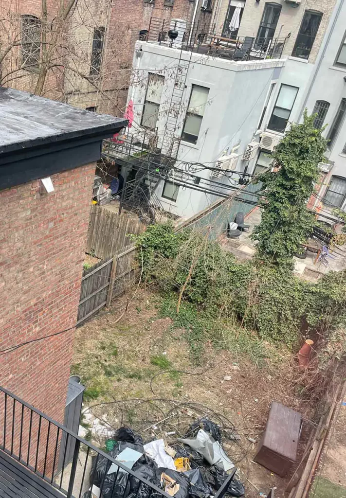 The overgrown Brooklyn backyard before the Real Simple and Decks R Us remodel. Garbage and piles of refuse are piled up in the backyard. A view from a balcony on the backside of the brownstone which displays the neighboring buildings.