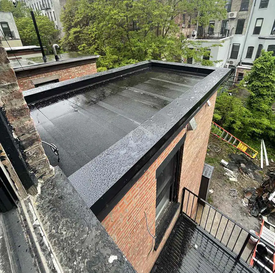 A before image of the rooftop at the Brooklyn brownstone before the deck remodel. It is painted black and appears to be an empty area covered with water puddles and droplets during a rainy day. The backyard is littered with a ladder, siding, and other construction waste.
