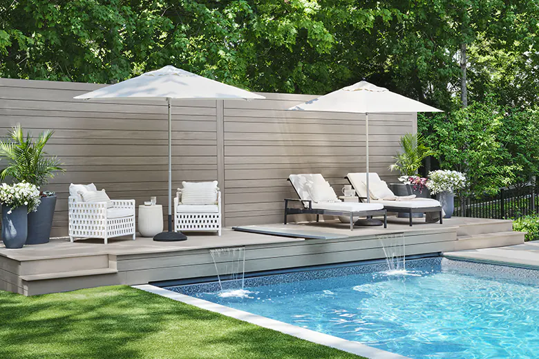 Side view of a backyard in-ground pool with a light gray deck and matching privacy wall. There are two sitting areas under umbrellas on the deck.