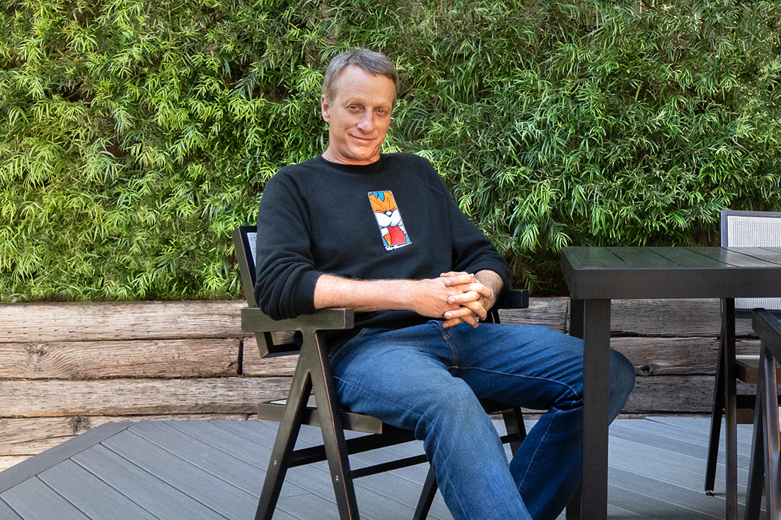Tony Hawk gently smiles with folded hands sitting in a chair on his new deck.