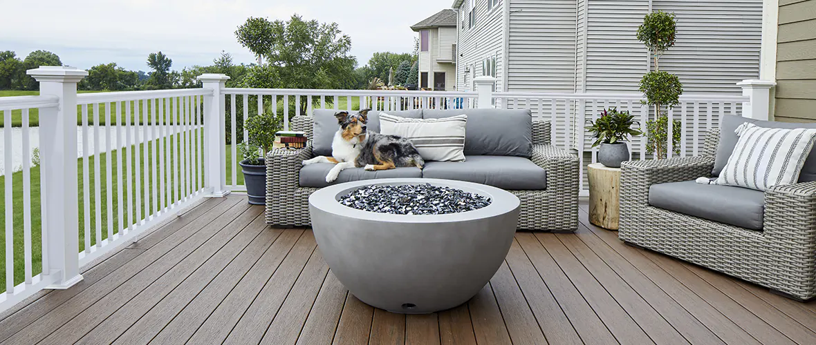 A dog lounges on outdoor furniture in front of a firepit on a deck.