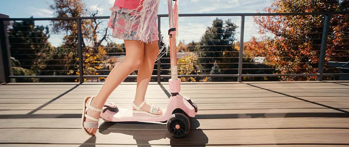 A child in open-toed shoes rides a scooter on a deck.