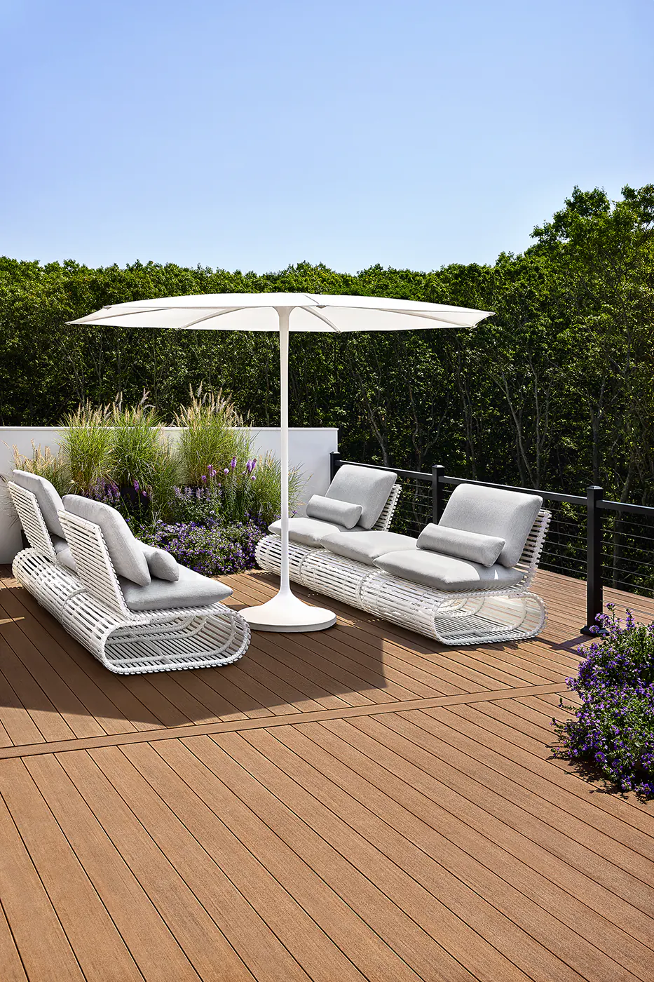Richly-hued reddish deck boards comprise an elevated deck. Modern outdoor sofas with bent wire feet face each other underneath an umbrella.