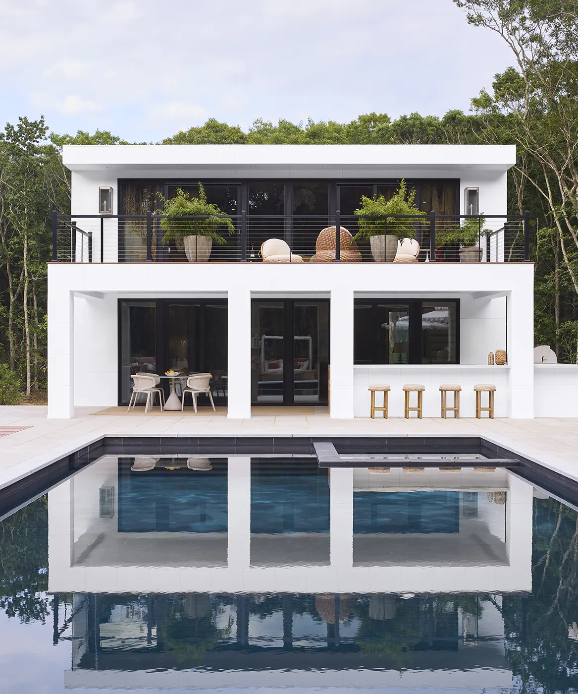 The back of the home features a second story deck with black horizontal cable wire infill railing. The picture is taken from the perspective of an expansive, deep blue in-ground pool in the backyard.